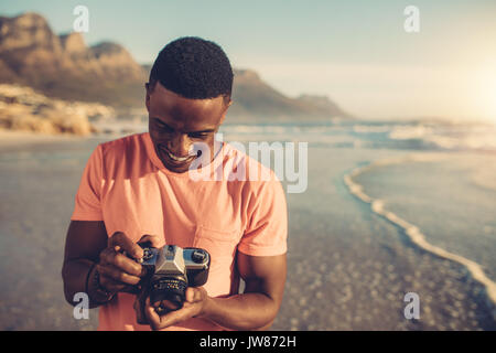 African man checking photos in his digital camera. Young man with digital photo camera standing on the beach. Stock Photo