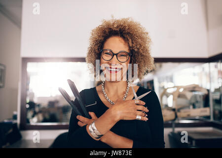 Hairdresser holding a hair straightener and scissors. Closeup of curly haired woman hairdresser in happy mood at the salon. Stock Photo