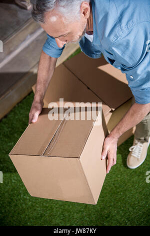 Senior man standing on green grass and holding big cardboard box Stock Photo