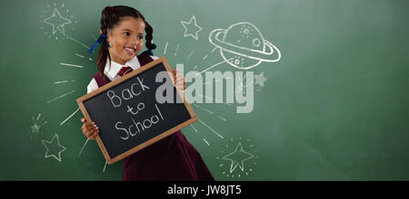 Smiling schoolgirl holding writing slate  against green chalkboard Stock Photo