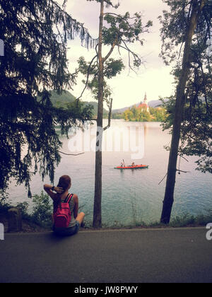 young women sitting on the shore of Lake Bled next to trees with view of church Stock Photo