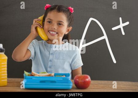 Digital composite of Happy student girl at table holding a banana against grey blackboard with A+ text Stock Photo