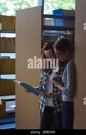 Female students reading book while standing by shelf in library Stock Photo