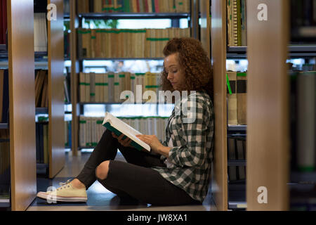 Side view of female teenage student reading book while sitting by shelf in library Stock Photo