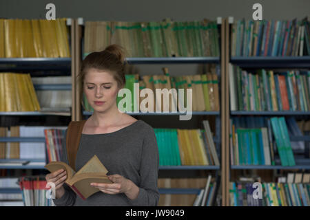 Young female student reading book while standing in library Stock Photo