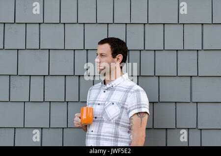 Side profile of an attractive man wearing a white plaid shirt holding an orange coffee cup against a grey background. Stock Photo