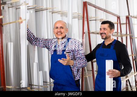 Two positive workmen in blue coverall with PVC windows and doors profiles at workshop. Selective focus Stock Photo