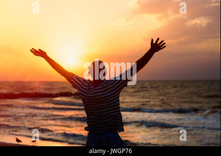 Silhouette of a man raising his hands to the sky at sunset on the beach and praying to God Stock Photo