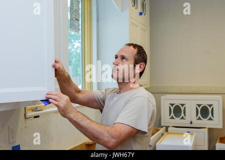 Repairman In Overalls Repairing Cabinet Hinge In Kitchen Installation of kitchen. Worker installs doors to kitchen cabinet. Stock Photo