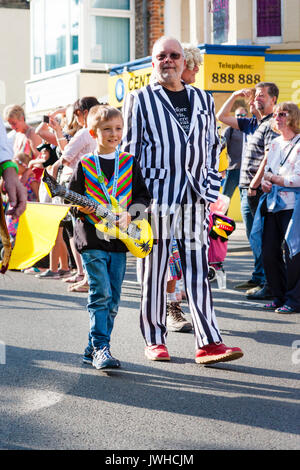 England, Broadstairs folk festival. The main parade. Mick Scott walking in black and white striped suit, young boy walking next to him with blow up guitar. Backlit. Stock Photo