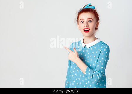 Excited young redhead caucasian girl with hair knot pointing her index finger sideways, raising eyebrows and keeping mouth wide opened, showing someth Stock Photo