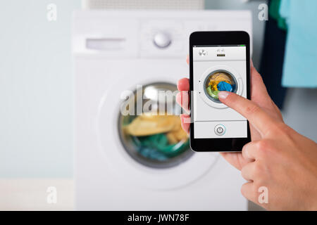 Close-up Of Person Hand Operating Washing Machine Using Mobile Phone Using App Stock Photo