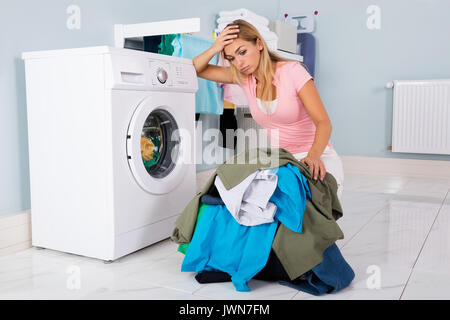 Unhappy Woman Showing Signs Of Fatigue Looking At Pile Of Clothes Near Washing Machine Stock Photo