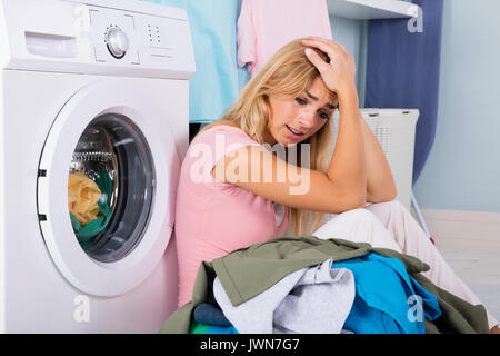 Unhappy Stressed Woman Showing Signs Of Fatigue Looking At Pile Of Clothes Near Washing Machine Stock Photo