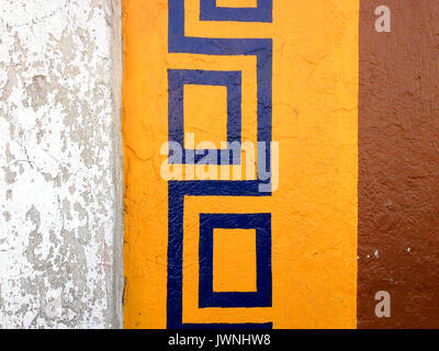 Colorful repeat pattern border painted on an exterior wall with a blue motif over a yellow frame with brown paint alongside, grungy section of wall to Stock Photo