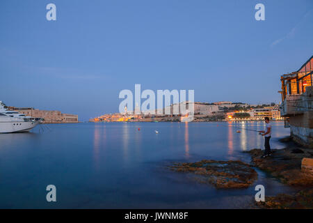 Summer night view of Valetta profile over sea. Long exposure. Illuminated architecture. Guy fishing on the shore. Stock Photo