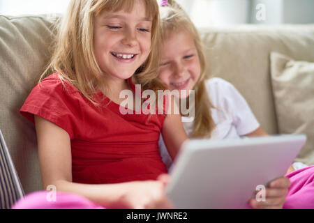 Cheerful little sisters gathered together in living room and watching their favorite cartoons on digital tablet Stock Photo