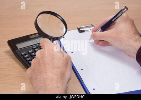 Man holding magnifying glass over black calculator and pen in fingers while writing something on blank note pad Stock Photo