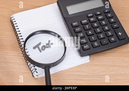 Calculation of taxes to be paid with a small magnifying glass highlighting the handwritten word - Tax - on a blank notepad with a calculator alongside Stock Photo