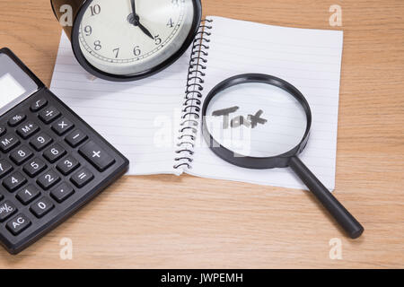 Close up of analogue clock, calculator and open notebook with the word tax highlighted by a magnifying glass on a timber office desk. Stock Photo
