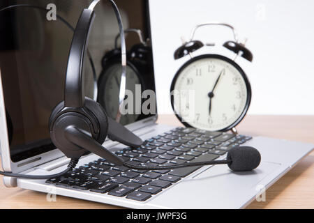 Alarm clock and headset on an open laptop computer with blank screen standing on a wooden desk n a concept of time management, deadlines and communica Stock Photo
