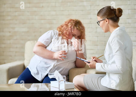 Portrait of  female psychiatrist  taking notes while consulting obese young woman crying with glass of water during therapy session on mental issues Stock Photo