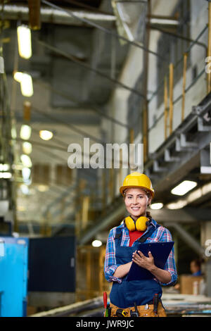 Portrait of young woman in hardhat working in modern plant, smiling at camera holding clipboard Stock Photo