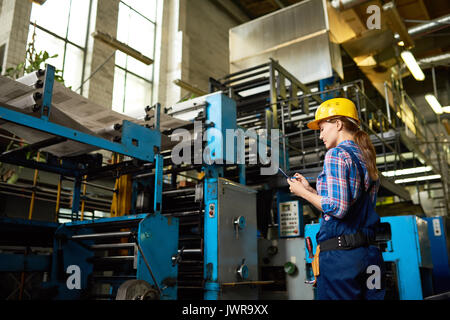 Portrait of female factory worker writing on clipboard checking operating machines with large printing unit in background Stock Photo
