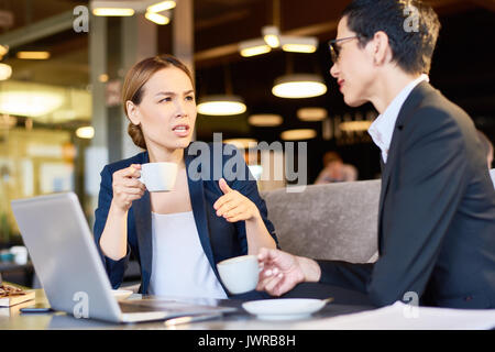 Multi-ethnic group of colleagues enjoying fragrant coffee while brainstorming on joint project, interior of spacious cafe on background Stock Photo