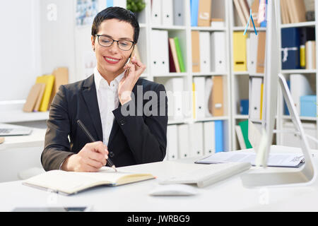 Attractive middle-aged designer writing down order details while talking to her client on mobile phone, interior of modern office on background Stock Photo
