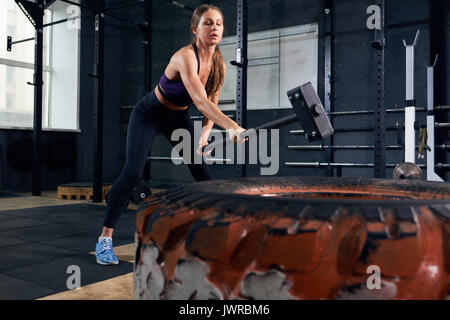 Portrait of strong young woman smashing large tire with sledgehammer during crossfit workout in modern gym Stock Photo