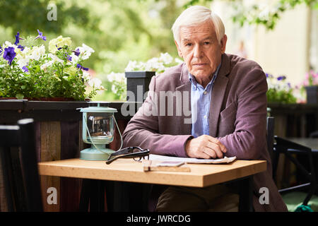 Portrait of thoughtful senior man sitting at table in outdoor cafe lounge with newspaper, looking at camera Stock Photo