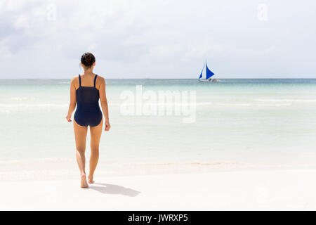 Young female in swimsuit walks out to the clear empty sea of Boracay beach with sailing boat on horizon. Stock Photo