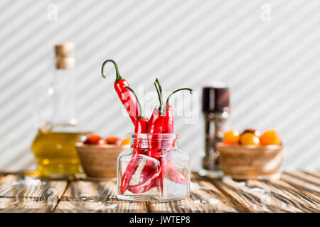 Red chili peppers in glass jar, sea salt and olive oil on wooden table. Harsh morning light Stock Photo