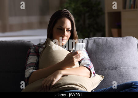 Sad girl holding pillow and reading phone message in the dark sitting on a couch at home Stock Photo