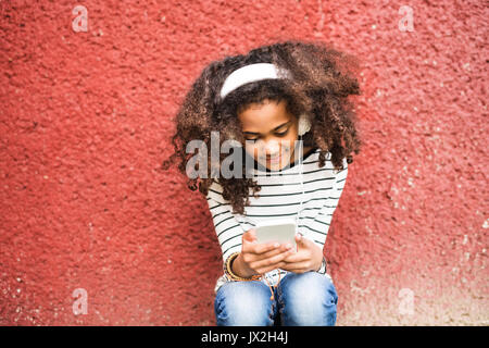 Beautiful african american girl with curly hair outdoors against red wall, holding smart phone, wearing yellow headphones, listening music. Stock Photo