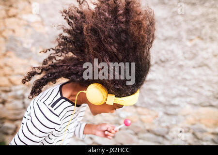Beautiful african american girl with curly hair outdoors, against stone wall, wearing headphones, listening music, eating lollipop. Stock Photo