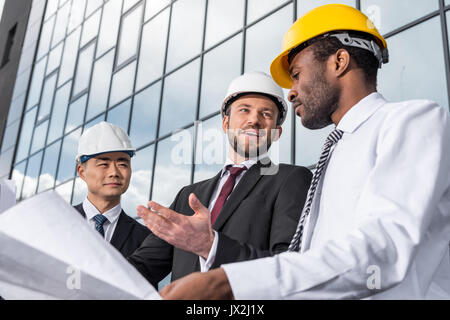Multiethnic group of professional architects in helmets working with blueprint outside modern building Stock Photo