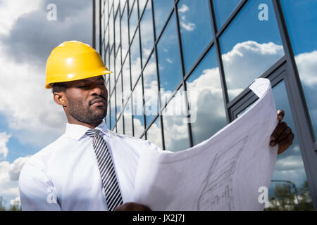 Low angle view of professional architect in hard hat holding blueprint outside modern building Stock Photo