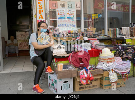 A Chinese American woman watches her shop while talking on the phone and fanning herself in Chinatown, Flushing, Queens, New York City Stock Photo
