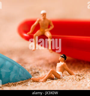 two different miniature men wearing swimsuit relaxing next to a blue plastic starfish and a red toy shovel on the sand of the beach Stock Photo