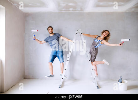 Beautiful young couple at home standing on ladder painting walls in their new house using paint rollers. Home makeover and renovation concept. Stock Photo