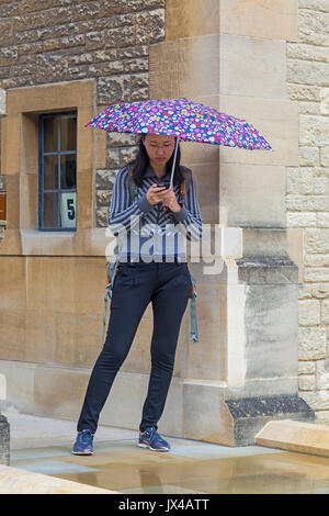 Woman using mobile phone sheltering under an umbrella on a wet rainy day at Oxford, Oxfordshire UK  in August