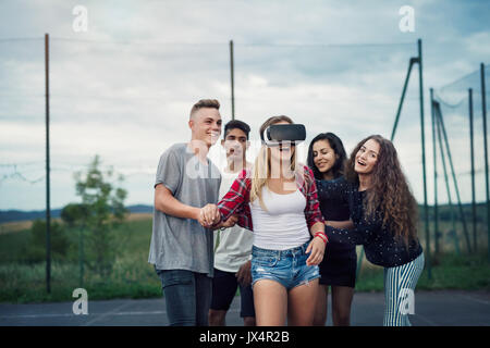 Group of attractive teenagers outdoors on playground, girl wearing virtual reality goggles. Stock Photo