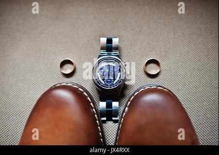 Close-up photo of groom's brown oxford shoes, a watch and two wedding rings. Stock Photo