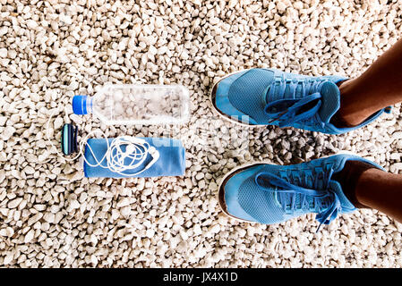 Feet of unrecognizable runner in blue sport shoes, water bottle, towel and earphones. Sports stuff on white gravel background. Stock Photo