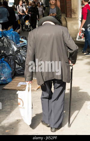 A hunched-up elderly man with a walking stick and carrier bag walking in central London, England, UK Stock Photo