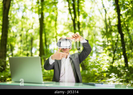 Confident handsome business man with virtual reality glasses pointed with hands at office desk in green park. Stock Photo