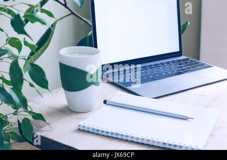 A modern office workplace surrounded by green plants. A wooden table with a laptop, notebook, pencil and a cup of water and lemon. Toned image Stock Photo