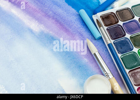aquarelle palette with paintbrushes and pastel crayons on blue watercolor background Stock Photo